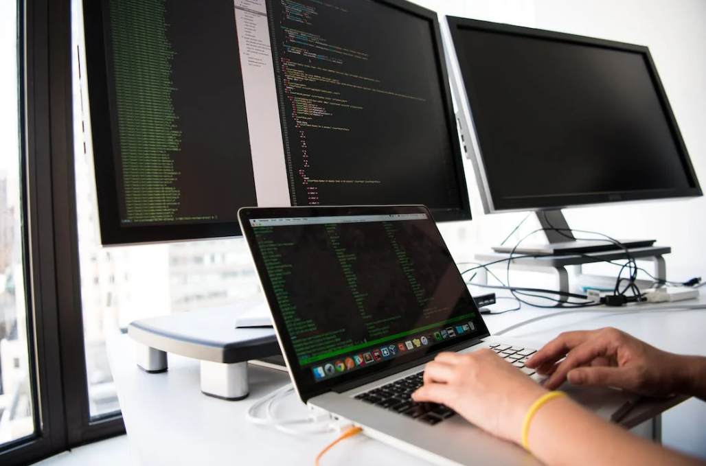 two computer screens on a white desk, and hands typing on the laptop with coding on the screen